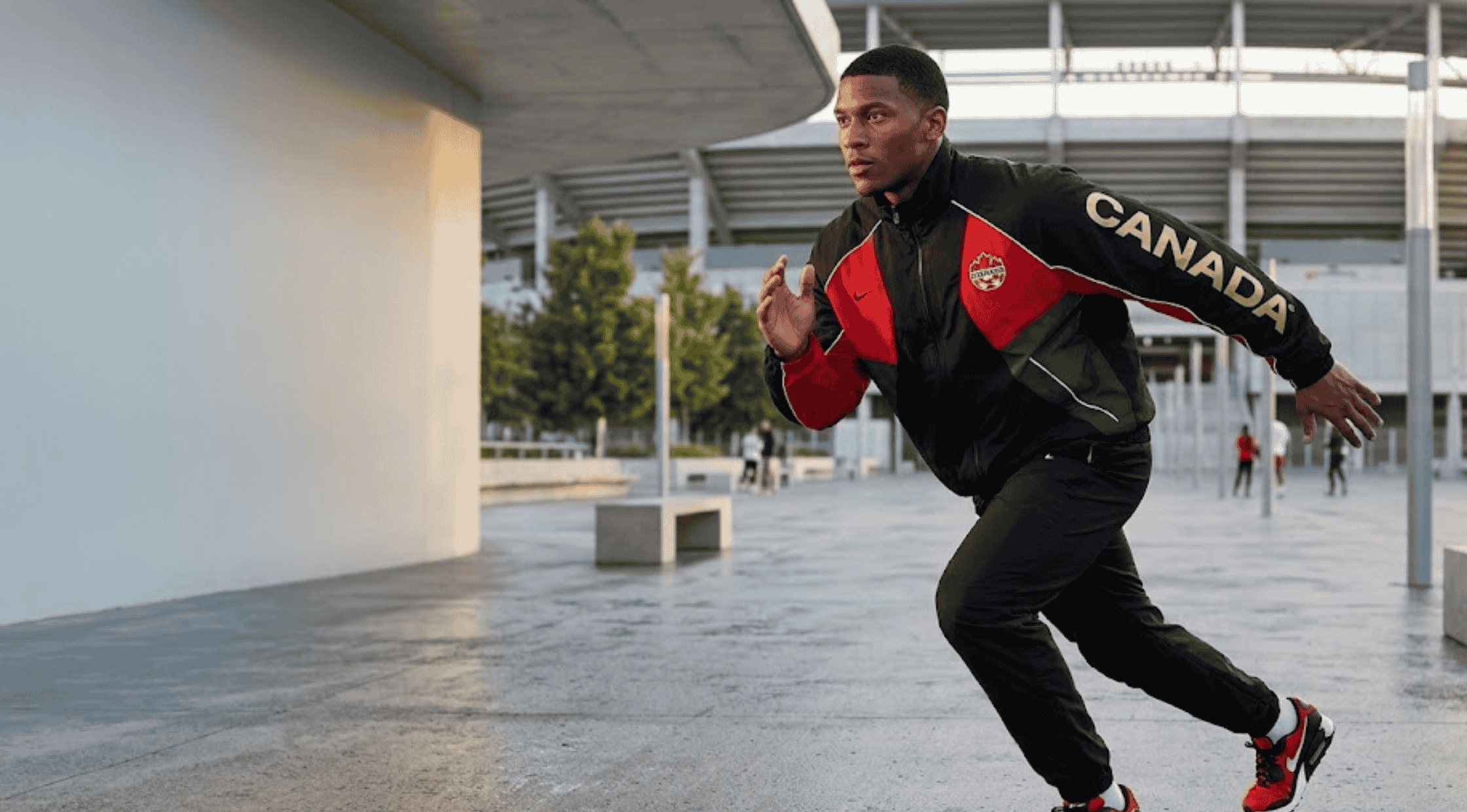 Athlete in a dynamic pose outside a stadium, wearing the NOCTA x Canada Soccer FIFA World Cup 2026 Track Jacket featuring red panels, gold Canada sleeve print, and the maple leaf crest.
